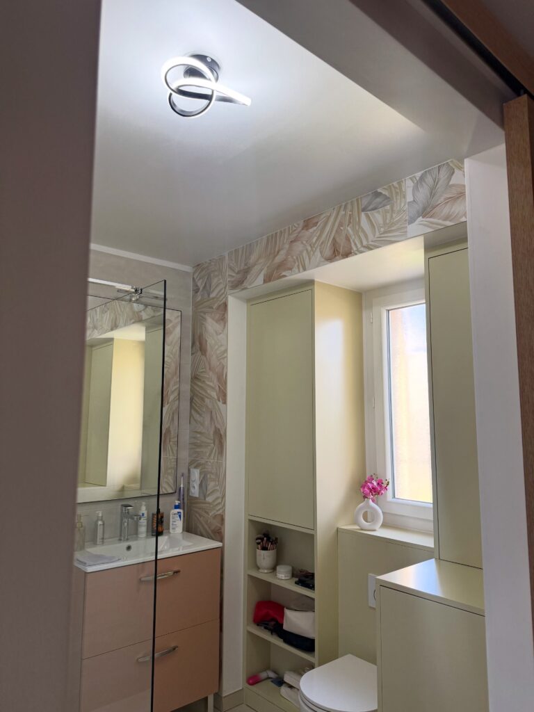 Salle de bain lumineuse avec meuble vasque beige, miroir et carrelage mural à motif feuillage.