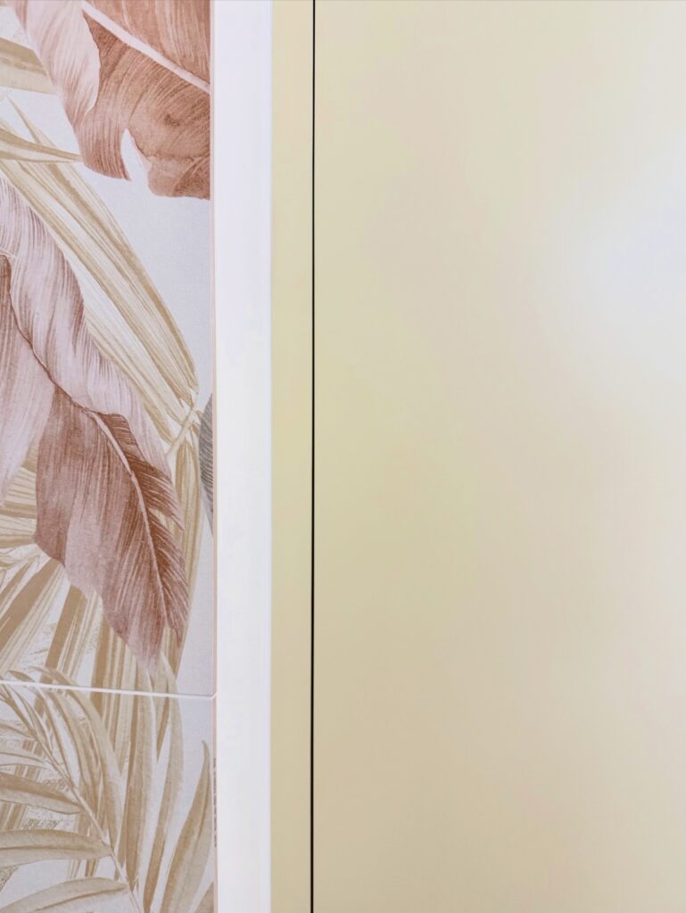 Tapisserie à motif feuilles de palmier beige et marron juxtaposé à un mur uni jaune pâle.