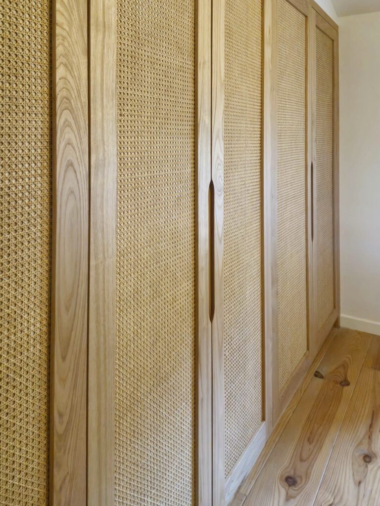 Placard en bois clair avec panneaux en cannage et poignées intégrées sur parquet.