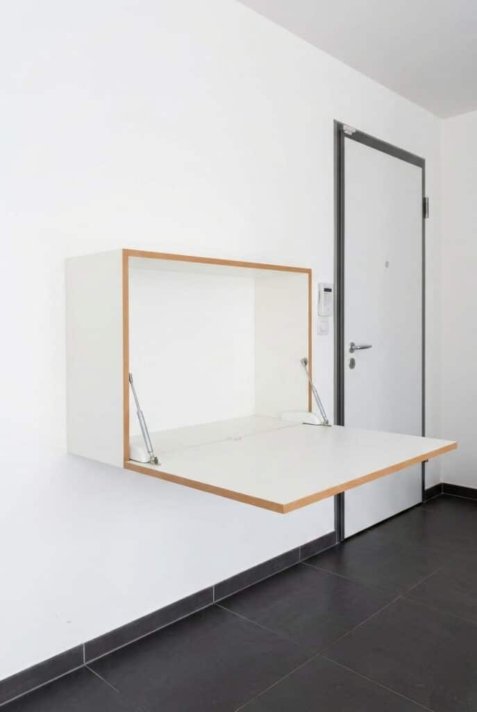 Bureau mural blanc escamotable avec bordures bois, ouvert à côté d'une porte sur mur blanc.