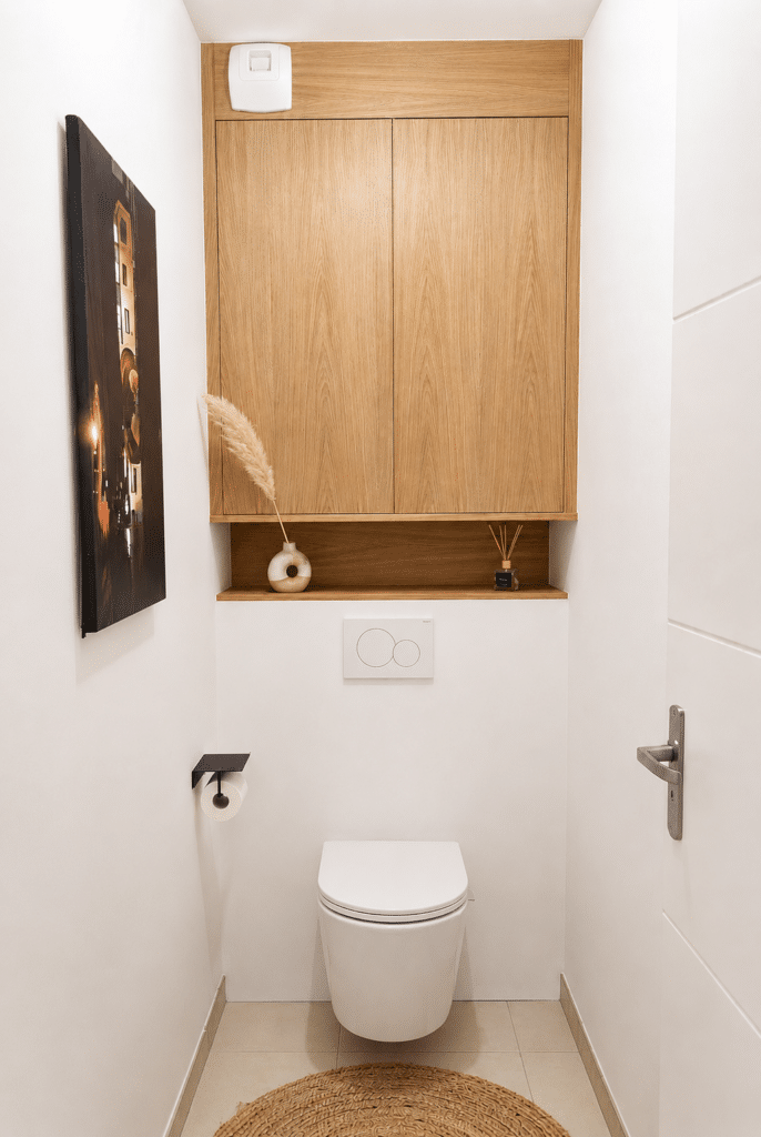 Toilettes suspendues blanches sous un placard en bois clair. Décoration épurée avec vase et tapis en jute.