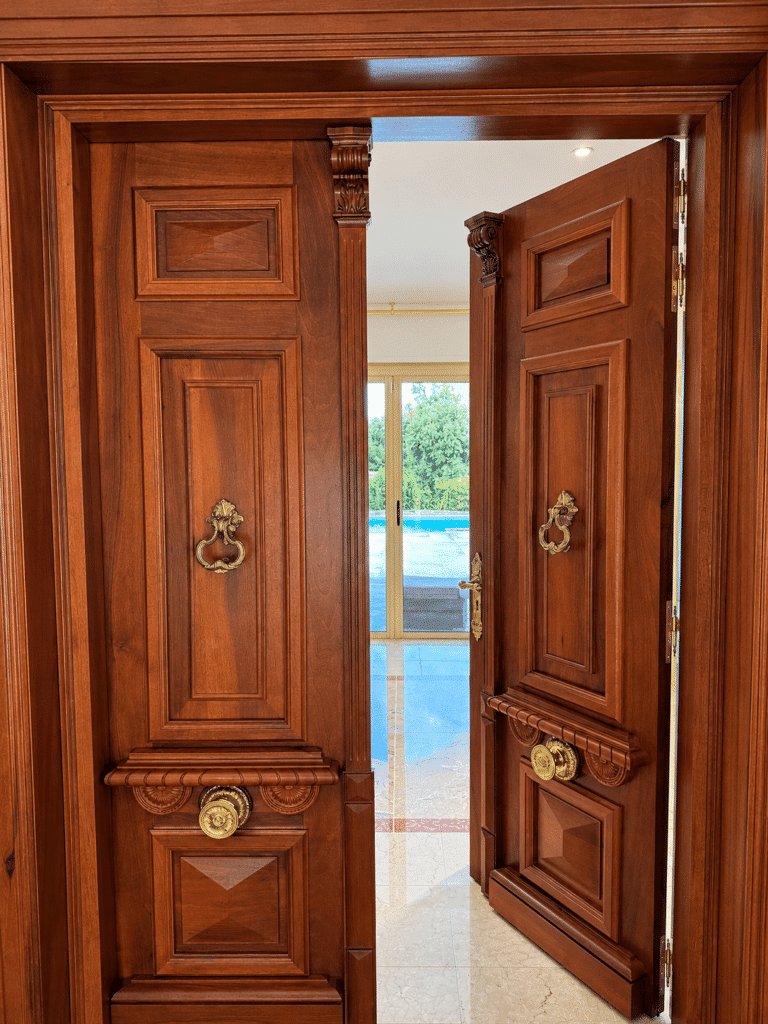 Double porte en bois noble sculpté avec heurtoirs en laiton doré ouvrant sur un intérieur luxueux avec piscine.