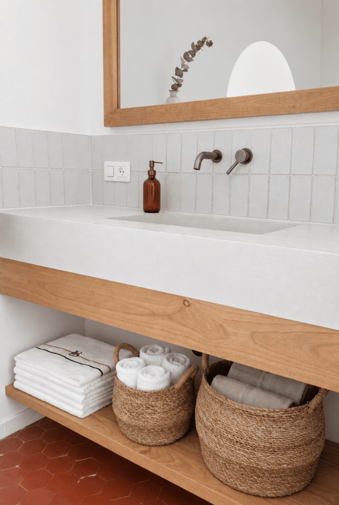 Salle de bain avec vasque blanche, meuble en bois, paniers en osier et robinetterie murale encastrée.
